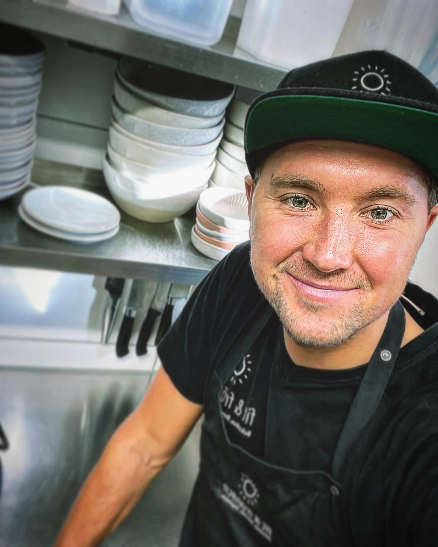 Geschäftsführer Tim Rühle von Fit und Fröhlich steht lächelnd in einer organisierten Restaurantküche, trägt eine schwarze Schürze und eine Kappe, umgeben von ordentlich gestapeltem Geschirr.