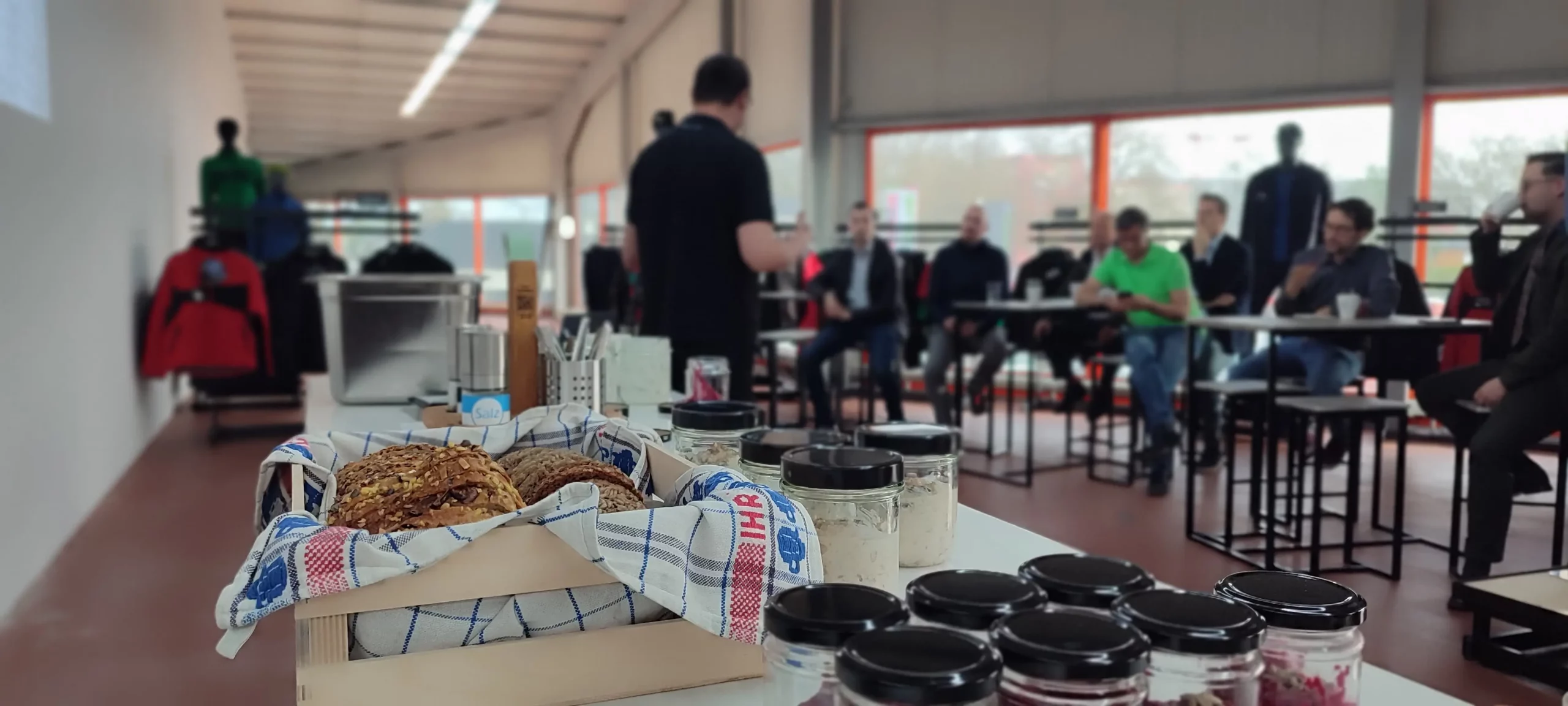 Unternehmer sitzen um Tische beim HR-Breakfast, während im Vordergrund frisch gebackenes Brot und Bircher Müsli angeboten werden, symbolisch für die Kombination von Innovation und menschlicher Verbindung.
